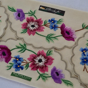 Könnte beinhalten: Cremefarbene Leinen-Tischsets mit gestickten Blumenmustern. Die Stickerei zeigt rosa, lila und blaue Blumen mit grünen Blättern. Die Tischsets haben einen gewellten Rand und den Text "All Pure Linen Made in Ireland".