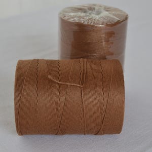 May include: Two spools of brown twine are displayed against a white background. One spool is in the foreground, unwrapped, and the other is in the background, wrapped in clear plastic. The twine is tightly wound, showing a textured surface.