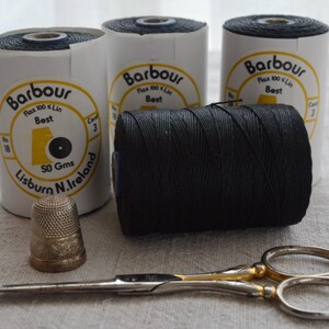 Puede incluir: Tres carretes de hilo de lino negro Barbour con etiquetas blancas que dicen 50 Gms y Lisburn N. Ireland, acompañados de un dedal metálico con superficie texturizada y unas tijeras de costura de metal plateado y dorado con dos anillas.