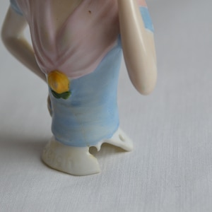 Pode incluir: Uma cabe&ccedil;a de boneca de porcelana com um vestido azul e rosa e um detalhe de flor amarela. A boneca est&aacute; faltando um peda&ccedil;o de sua base.