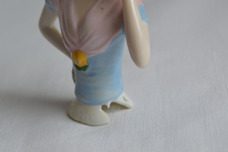 Pode incluir: Uma cabe&ccedil;a de boneca de porcelana com um vestido azul e rosa e um detalhe de flor amarela. A boneca est&aacute; faltando um peda&ccedil;o de sua base.