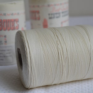 Peut inclure: Gros plan sur une bobine de fil blanc cassé à surface texturée. Deux autres bobines sont légèrement floues en arrière-plan. Les bobines portent des étiquettes avec du texte, dont la marque "BARBOUR'S".