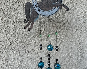 May include: A rustic metal wind chime featuring a horse silhouette inside a horseshoe. The wind chime has turquoise and black beads, and brown bell-shaped accents. The horse and horseshoe are painted with a distressed finish, adding to the charm.