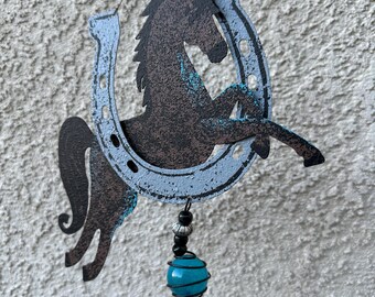 May include: A rustic metal wind chime featuring a horse silhouette within a horseshoe. The horse and horseshoe have a distressed, textured finish with blue accents. A turquoise glass bead and a wooden bell dangle below, suspended by a wire.