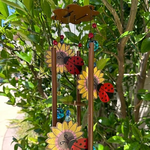 May include: A decorative wind chime featuring sunflowers and ladybugs. The wind chime has brown metal tubes, a brown top with a cloud shape, and colorful beads. The sunflowers are yellow with brown centers, and the ladybugs are red with black spots.