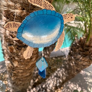 May include: A blue and brown wooden turtle wind chime with a white sandy beach design and blue glass beads hanging from it.