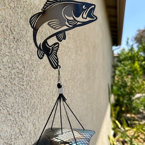 May include: Black metal wind chime featuring a silhouette of a bass fish jumping out of the water. The chime has a hammered metal top and black cords.