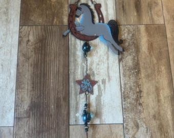 May include: A rustic metal wind chime featuring a gray horse leaping over a horseshoe. Below, a star with turquoise accents, blue beads, and a brown cylindrical piece hang from a chain. The wind chime is designed with a Western theme.