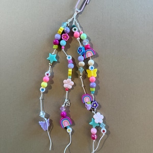 May include: A beaded charm with a paperclip attachment. The charm features colorful beads, stars, flowers, butterflies, and rainbows. The beads are in shades of pink, blue, yellow, and green. The charm is attached to a light purple paperclip.