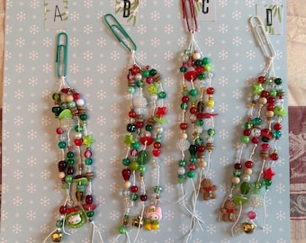 May include: Four beaded Christmas ornaments with paperclip attachments. Each ornament features a variety of beads in red, green, white, and brown, with small charms and bells. The ornaments are displayed on a light blue background with snowflake patterns.