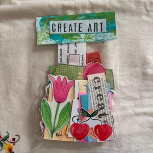 May include: A clear plastic bag filled with colorful paper ephemera. The bag includes a pink tulip, red cherries, and various paper scraps. A banner at the top reads "CREATE ART".
