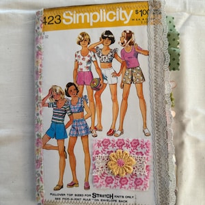 May include: A vintage Simplicity sewing pattern book, featuring illustrations of women's tops and shorts. The cover is decorated with a pink floral fabric and a gold flower embellishment. The text "423 Simplicity" is visible.
