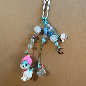 May include: A whimsical paperclip charm featuring a white and blue cat figurine wearing a pink turban and red collar. The charm includes various beads in shades of blue, purple, and white, along with a small mouse, a tooth, and a crescent moon.