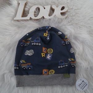 Könnte beinhalten: Eine blaue Strickmütze mit grauem Rand. Die Mütze hat ein Muster aus gelben Bauwagen und Blöcken mit den Buchstaben "WORK" darauf.