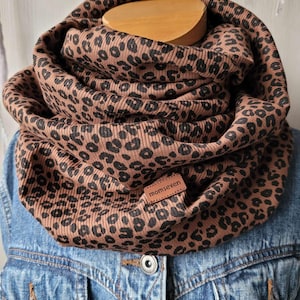 Könnte beinhalten: Ein braun-schwarzer Loop-Schal mit Leopardenmuster. Der Schal ist aus geripptem Stoff gefertigt und hat ein kleines Lederetikett mit der Aufschrift "momseven". Der Schal ist um eine Holzform gewickelt und wird auf einer Jeansjacke präsentiert.