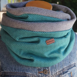 Könnte beinhalten: Ein türkisfarbener und grauer Loop-Schal mit Fischgrätenmuster. Der Schal hat ein graues Fleecefutter und ein kleines braunes Lederetikett mit der Aufschrift "momseven". Der Schal wird auf einer Jeansjacke präsentiert.