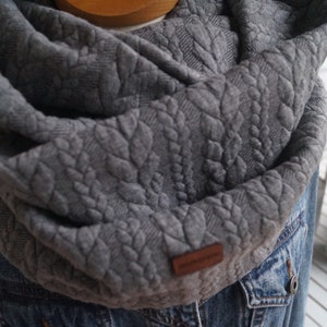 Könnte beinhalten: Grauer gestrickter Infinity-Schal mit einem strukturierten Muster. Der Schal ist gefaltet und über eine Jeansjacke gelegt.