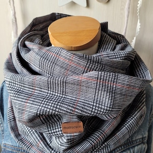 Könnte beinhalten: Ein schwarzer und weißer karierter Infinity-Schal mit einem roten Faden, der durch ihn verläuft. Der Schal befindet sich auf einer Holzpuppe und hat ein braunes Leder-Etikett mit dem Text "momseven" darauf.