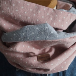 Könnte beinhalten: Ein rosa-grauer Loop-Schal mit weißen Punkten. Der Schal ist aus weichem Stoff gefertigt und hat ein Wendemotiv. Ein kleines braunes Etikett mit dem Wort "momseven" ist auf den Schal genäht.