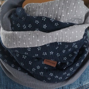 Könnte beinhalten: Ein grauer und marineblauer Infinity-Schal mit weißem Blumenmuster. Der Schal hat ein kleines braunes Etikett mit dem Text "momskreativ".
