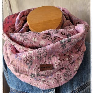 Könnte beinhalten: Ein rosa Infinity-Schal mit Blumenmuster und einem braunen Lederetikett mit der Aufschrift "momseven". Der Schal ist um eine Holzform gewickelt und vor einer Jeansjacke platziert. Der Schal hat ein zartes Blumenmuster.