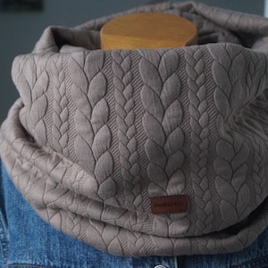 Könnte beinhalten: Ein grauer gestrickter Infinity-Schal mit einem Zopfmuster. Der Schal hat ein braunes Leder-Etikett mit dem Wort "moonriver" darauf.