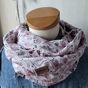 Könnte beinhalten: Ein Schal mit floralem Muster in Rosa-, Lila- und Weißtönen, der auf einer Schaufensterpuppe präsentiert wird. Der Schal hat ein sich wiederholendes Muster aus Blumen und Blättern. Ein kleines braunes Lederetikett mit dem Text "momseven" ist auf den Schal genäht.
