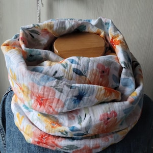 Könnte beinhalten: Ein weißer Loop-Schal mit einem Blumenmuster in Orange, Rosa, Blau und Gelb. Der Schal besteht aus einem weichen, strukturierten Stoff und ist um eine Holzform gewickelt. Der Schal wird über einer Jeansjacke getragen.