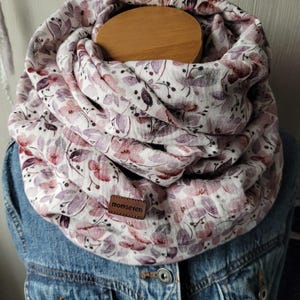 Könnte beinhalten: Ein Schal mit floralem Muster in Rosa, Lila und Weiß. Der Schal ist um eine Holzform gewickelt und über einer Jeansjacke getragen. Ein kleines Lederetikett mit dem Text "momseven" ist angebracht.