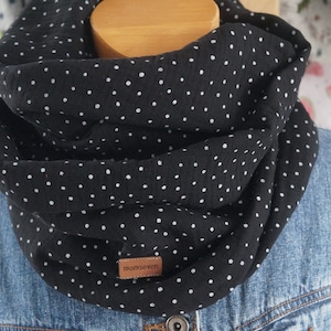 Könnte beinhalten: Schwarzer Loop-Schal mit weißen Punkten. Der Schal ist um eine Holzform und eine Jeansjacke gelegt. Ein kleines braunes Lederetikett mit dem Text "momseven" ist sichtbar.