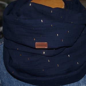 Könnte beinhalten: Ein marineblauer Loop-Schal mit goldenen Metallic-Akzenten. Der Schal ist aus einem weichen Stoff gefertigt und hat ein kleines braunes Lederetikett mit dem Text "momseven". Der Schal ist um den Hals gewickelt.