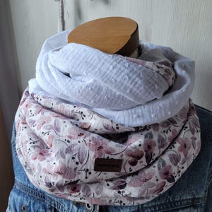 Könnte beinhalten: Ein mehrlagiger Loop-Schal mit einem floralen Muster in Rosa- und Lilatönen, kombiniert mit einem weißen, strukturierten Stoff. Der Schal wird auf einem hölzernen Mannequinkopf über einer Jeansjacke präsentiert.