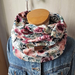 Könnte beinhalten: Ein Schal mit floralem Muster in Burgunder-, Rosa- und Grüntönen auf weißem Hintergrund. Der Schal ist um eine Schaufensterpuppe gewickelt und hat ein kleines braunes Lederetikett mit der Aufschrift "momseven". Der Schal wird über einer Jeansjacke getragen.