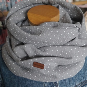 Könnte beinhalten: Grauer Loop-Schal mit weißen Polka-Dots, drapiert um eine Holzform. Der Schal besteht aus einem weichen, strukturierten Stoff. Ein kleines braunes Lederetikett mit dem Wort "momseven" ist auf den Schal genäht.