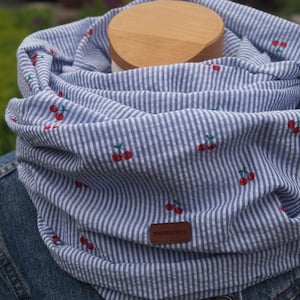 Könnte beinhalten: Ein blauer und weiß gestreifter Infinity-Schal mit roter Kirschen-Stickerei. Der Schal hat ein braunes Leder-Etikett mit dem Wort "momseven" darauf.