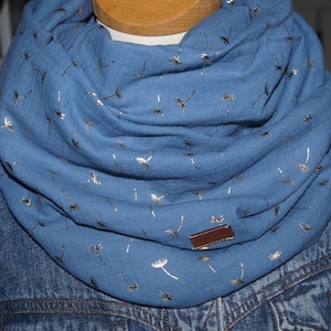 Könnte beinhalten: Blauer Loop-Schal mit goldenem Pusteblumenmuster. Der Schal ist um den Hals einer Schaufensterpuppe gelegt und wird mit einer Jeansjacke kombiniert. Ein kleines braunes Lederetikett ist am Schal befestigt.