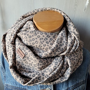 Könnte beinhalten: Ein beigefarbener und grauer Schal mit Leopardenmuster. Der Schal besteht aus einem gerippten Stoff und hat ein kleines braunes Lederetikett mit der Aufschrift "momseven". Der Schal ist über einer Holzform und einer Jeansjacke drapiert.