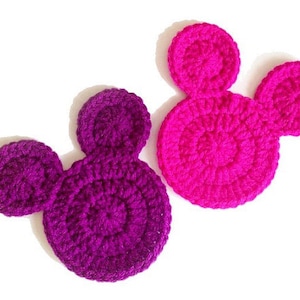 May include: Two crocheted appliques in the shape of a mouse head. One is a deep purple, the other is a vibrant pink. The appliques are made with a textured yarn and have a circular design.