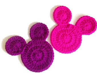 May include: Two crocheted appliques in the shape of a mouse head. One is a deep purple, the other is a vibrant pink. The appliques are made with a textured yarn and have a circular design.