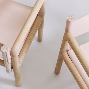 May include: Two wooden chairs with light pink leather seats. The chairs have a simple, minimalist design with rounded edges.