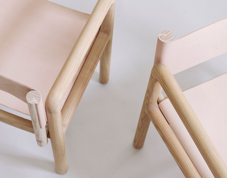 May include: Two wooden chairs with light pink leather seats. The chairs have a simple, minimalist design with rounded edges.