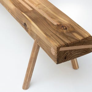 Puede incluir: Un banco de madera con un acabado natural y un diseño único. El banco tiene un asiento ancho y rectangular y cuatro patas de madera. El asiento está hecho de varias piezas de madera que se unen, creando un aspecto rústico. Las patas también están hechas de varias piezas de madera que se unen, creando un diseño único e interesante.