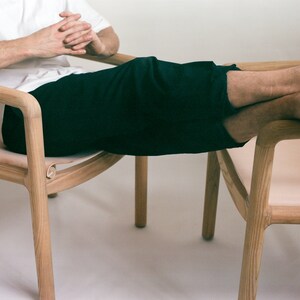 May include: A person sits in a wooden chair with a light-colored leather seat. The chair has a curved back and arms. The person's legs are resting on a second chair with a similar design.