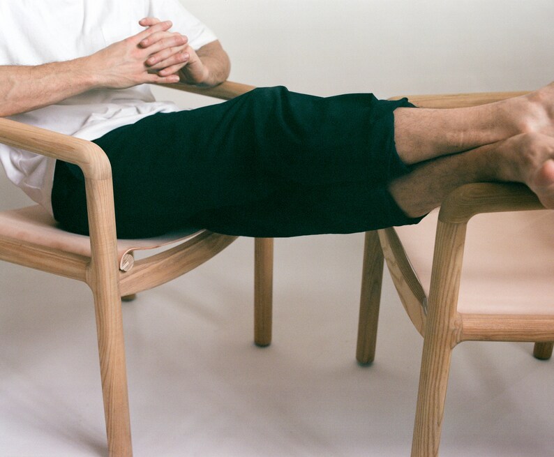 May include: A person sits in a wooden chair with a light-colored leather seat. The chair has a curved back and arms. The person's legs are resting on a second chair with a similar design.