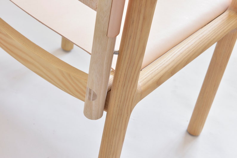 May include: Close-up of a wooden chair with a light brown finish. The chair has a curved back and a simple design. The chair is made of solid wood and has a natural grain pattern.