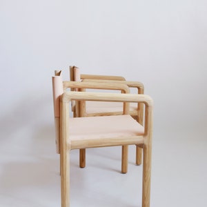 May include: Three wooden chairs with light pink leather seats and backs. The chairs are arranged in a row, with the backs of the chairs facing the camera. The chairs are made of light wood and have a simple, modern design.
