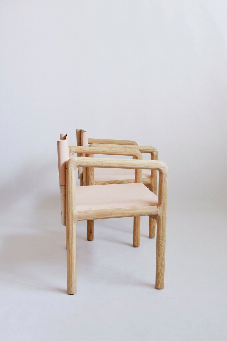 May include: Three wooden chairs with light pink leather seats and backs. The chairs are arranged in a row, with the backs of the chairs facing the camera. The chairs are made of light wood and have a simple, modern design.