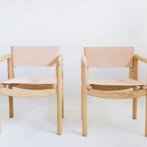 May include: Two wooden chairs with light pink leather seats and backs. The chairs have a simple, modern design with rounded edges.