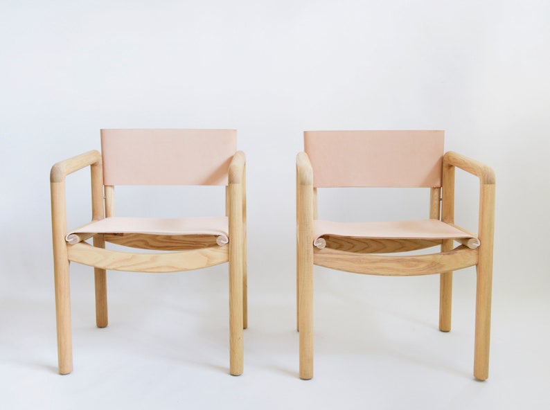 May include: Two wooden chairs with light pink leather seats and backs. The chairs have a simple, modern design with rounded edges.