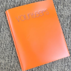 May include: A bright orange notebook with a smooth, matte cover. The word "volunteers" is embroidered in dark blue thread across the upper center of the front cover in a lowercase, rounded, sans-serif font style with visible stitching.
